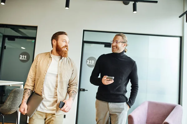 Happy bearded and tattooed man with laptop and smartphone talking to colleague in eyeglasses and black turtleneck in modern office with high tech interior, productivity and cooperation concept — Stock Photo