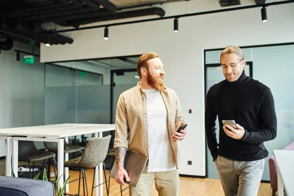 Cheerful, bearded and tattooed businessman with laptop talking to smiling colleague looking at mobile phone while standing in modern office, productivity and cooperation concept — Stock Photo