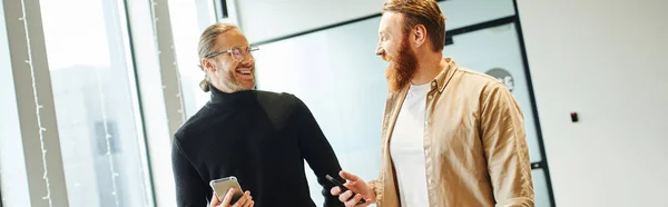 Pleased and successful business partners holding mobile phones while discussing startup planning in contemporary coworking office, productivity and cooperation concept, banner — Stock Photo