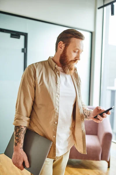Hombre de negocios barbudo y tatuado con ropa casual elegante que sostiene el ordenador portátil y la red en el teléfono móvil mientras está de pie en la oficina moderna, el concepto de emprendimiento exitoso - foto de stock