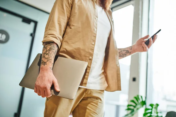 Partial view of tattooed entrepreneur in stylish casual clothes standing with laptop and messaging on mobile phone in modern office environment, successful business concept — Stock Photo