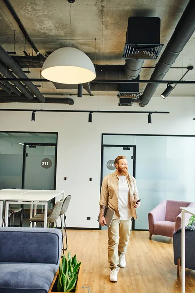 Full length of bearded, tattooed and stylish businessman in casual clothes walking with laptop and smartphone in modern office with high tech interior, successful entrepreneurship concept — Stock Photo