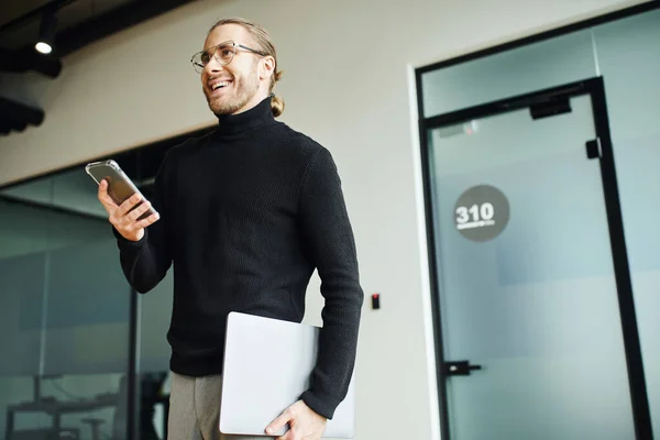 Homem de negócios elegante e ambicioso em óculos e gola alta preta de pé com smartphone e laptop, sorrindo e olhando para longe no escritório moderno, conceito de negócio bem sucedido — Fotografia de Stock