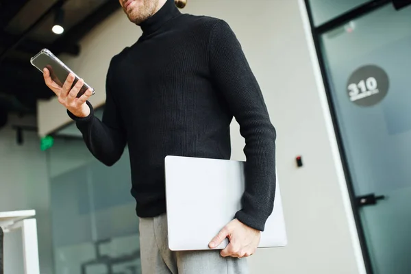 Partial view of entrepreneur in black turtleneck holding laptop and networking on mobile phone while standing in modern coworking office, successful business concept — Stock Photo