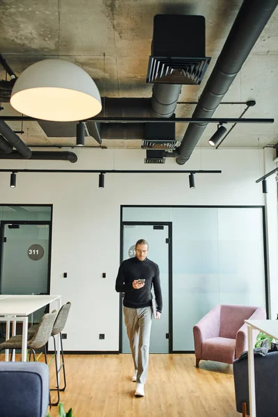 Полная длина стильный бизнесмен в очках и черная водолазка сообщения на смартфоне и прогулки в гостиной современного офиса с высокотехнологичным интерьером, успешная бизнес-концепция — стоковое фото