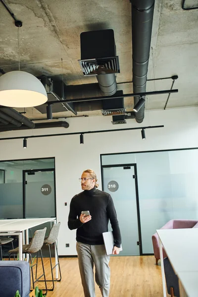 Career oriented businessman in black turtleneck and stylish eyeglasses walking with laptop and mobile phone while looking away in modern coworking space, successful business concept — Stock Photo