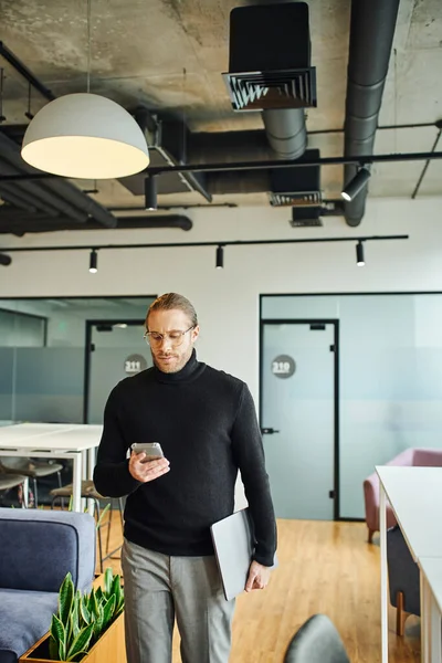 Serious and stylish entrepreneur in eyeglasses and black turtleneck holding laptop and networking on smartphone in contemporary office with high tech interior, successful business concept — Stock Photo