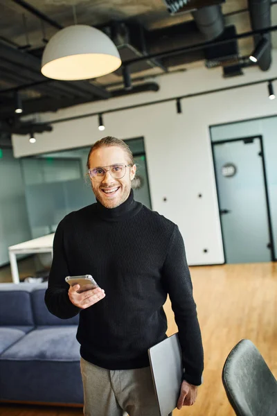 Professional headshot of cheerful businessman in black turtleneck holding laptop and smartphone while looking at camera in modern office with high tech interior, successful business concept — Stock Photo