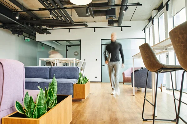 Motion blur of businessman holding laptop and walking in contemporary lounge of coworking space with modern furniture and green plants, full length, successful business concept — Stock Photo