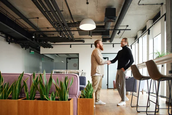 Side view of tattooed and bearded entrepreneur shaking hands with stylish colleague while confirming agreement in office lounge with modern furniture, high tech interior and natural plants — Stock Photo