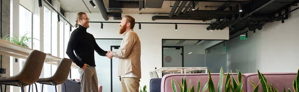 Side view of entrepreneur in stylish casual clothes shaking hands with bearded and tattooed business partner and closing deal in contemporary coworking office with high tech interior, banner — Stock Photo