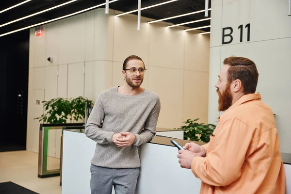 Excited businessman in eyeglasses and casual clothes talking to bearded colleague at reception in lobby of contemporary coworking office, entrepreneurs discussing possibility of startup — Stock Photo