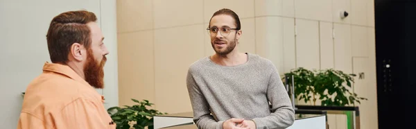 Inspired entrepreneur in casual clothes and eyeglasses listening to bearded business partner talking about startup project in lobby of modern office environment, creative thinking, banner — Stock Photo