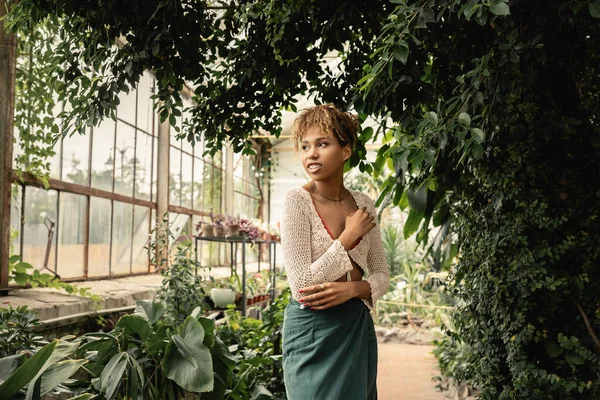 Sorrindo e na moda jovem mulher afro-americana no verão tricotado top e saia olhando para longe enquanto estava perto de plantas verdes no centro do jardim, mulher elegante desfrutando de um ambiente tropical exuberante — Fotografia de Stock