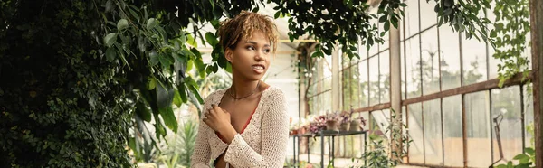 Sorrindo jovem afro-americana no verão top de malha olhando para longe, enquanto está perto de plantas verdes no centro do jardim, mulher elegante desfrutando de um ambiente tropical exuberante, banner — Fotografia de Stock