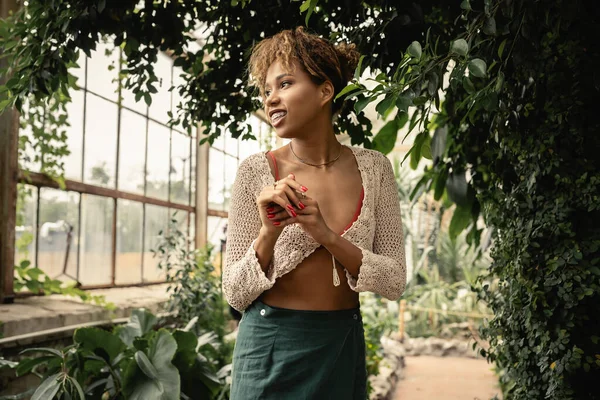 Positif jeune femme afro-américaine à la mode été tricoté haut et jupe détournant les yeux tout en se tenant près de plantes vertes dans le centre de jardin, femme élégante bénéficiant d'un environnement tropical luxuriant — Photo de stock