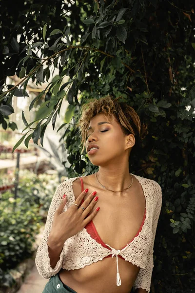 Portrait de jeune femme afro-américaine à la mode avec un maquillage en tricot tout en se tenant les yeux fermés près des plantes dans le centre de jardin, femme élégante bénéficiant d'un environnement tropical luxuriant — Photo de stock