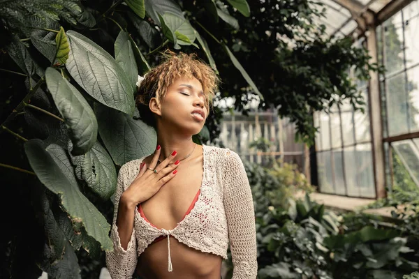 Portrait de la jeune femme afro-américaine élégante en tricot haut toucher cou tout en se tenant près du feuillage vert de plantes dans une serre floue, dame à la mode dans l'oasis tropicale — Photo de stock