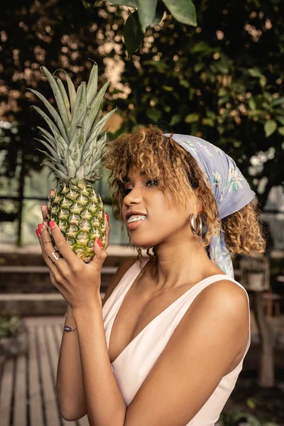 Ritratto di donna afroamericana sorridente ed elegante con bretelle in fazzoletto che regge l'ananas fresco e distoglie lo sguardo mentre si trova in una serra sfocata, donna alla moda in un'oasi tropicale — Foto stock