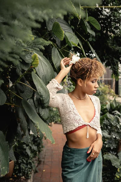 Mujer afroamericana joven de moda en la parte superior de punto y falda tocando el cabello y de pie cerca de follaje verde en el jardín interior borroso, señora de moda en oasis tropical - foto de stock