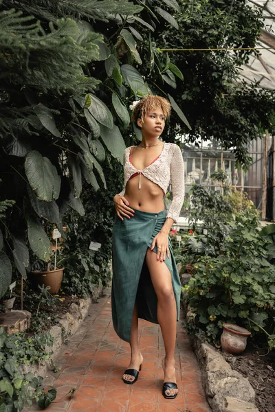 Full length of trendy young african american woman in skirt and knitted top looking away and standing near green plants in greenhouse, stylish lady surrounded by lush greenery, summer — Stock Photo