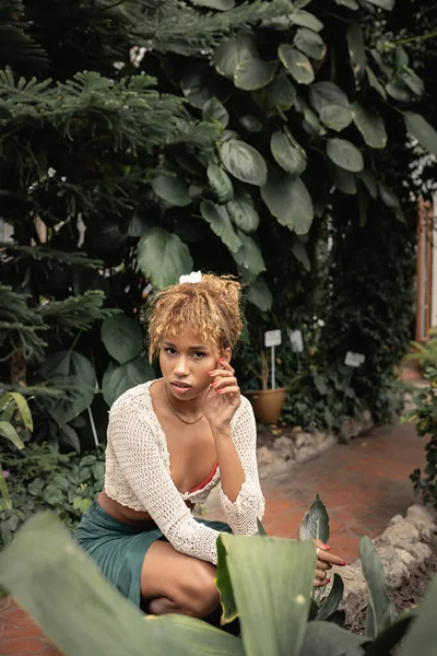 Alla moda giovane donna africana americana in abito estivo guardando la macchina fotografica mentre posa vicino a piante tropicali sullo sfondo in aranciata, signora elegante circondata da verde lussureggiante, estate — Foto stock