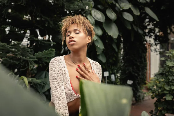 Mujer afroamericana joven y elegante en traje de punto de verano tocando el pecho y cerrando los ojos mientras está de pie en el jardín interior borroso en el fondo, fashionista mezclándose con la flora tropical — Stock Photo