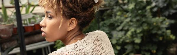 Vue latérale de la jeune femme afro-américaine en été haut tricoté regardant loin tout en passant du temps dans le centre de jardin flou à l'arrière-plan, fashionista se mélangeant avec la flore tropicale, bannière — Photo de stock