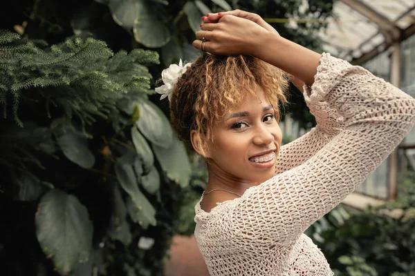 Porträt einer lächelnden jungen afrikanisch-amerikanischen Frau mit Hosenträgern und sommerlichem Outfit, die in die Kamera blickt, während sie neben grünen Pflanzen im Garten steht und sich mit tropischer Flora vermischt — Stockfoto