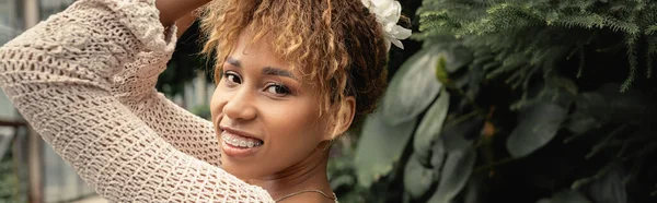 Femme afro-américaine à la mode et souriante avec des bretelles posant en haut tricoté d'été et regardant la caméra près de plantes vertes en orangerie, fashionista se mélangeant à la flore tropicale, bannière — Photo de stock