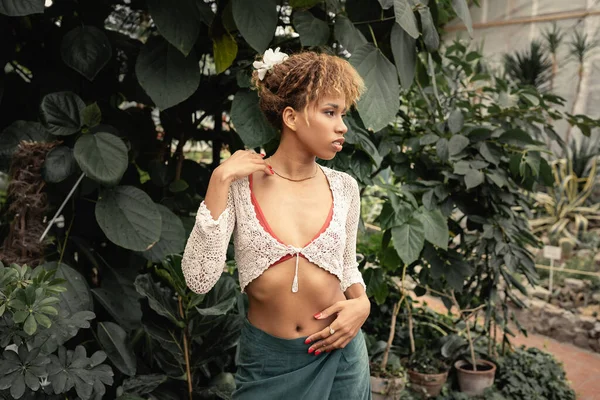 Стильная молодая африканская женщина в летнем наряде, отводящая взгляд, стоя рядом с зелеными растениями на заднем плане в садовом центре, модница смешивается с растительностью — стоковое фото