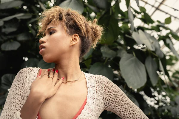 Vue à angle bas de la jeune femme afro-américaine à la mode en tricot haut d'été touchant l'épaule et regardant loin tout en se tenant près du feuillage vert, fashionista mélange avec la flore tropicale — Photo de stock