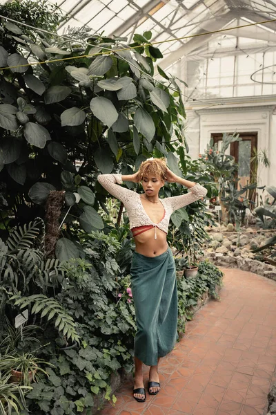 Full length of trendy young african american woman in knitted top and skirt touching head and standing near plants in orangery at background, fashionista blending in with tropical flora — Stock Photo