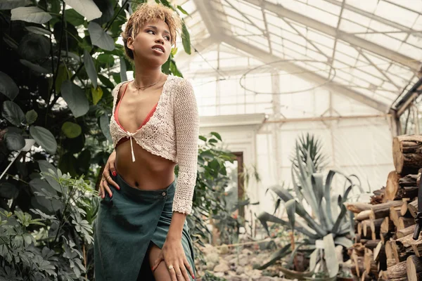 Modische junge Afroamerikanerin im Sommeroutfit, die die Hüfte berührt und wegschaut, während sie im verschwommenen Gartencenter steht, stilvolle Frau mit tropischem Hintergrund — Stockfoto