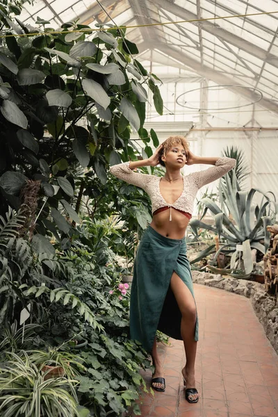 Full length of trendy young african american woman in summer knitted top and skirt standing and posing near green foliage in greenhouse, stylish woman with tropical backdrop — Stock Photo