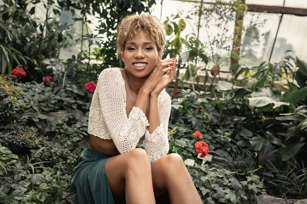 Jovem mulher americana africana alegre em roupa de verão e top de malha olhando para a câmera enquanto se senta perto de plantas borradas com flores em orangery, senhora moda-forward em meio a vegetação tropical — Fotografia de Stock