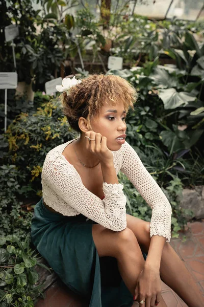 Portrait de jeune femme afro-américaine élégante en tenue d'été relaxante près de plantes vertes en arrière-plan dans une serre floue, dame avant-gardiste au milieu de la verdure tropicale — Photo de stock