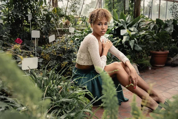 Молодая африканская американка в летнем наряде и вязаном топе, смотрящая в камеру, проводя время рядом с растениями на заднем плане в центре сада, модная леди среди тропической зелени — стоковое фото