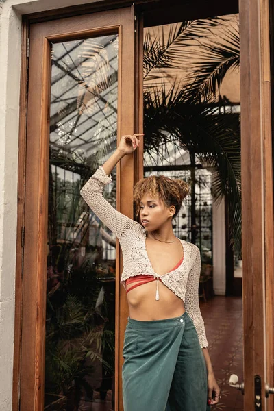 Молода афроамериканка в стильному літньому вбранні і в'язана зверху стоїть біля дверей садового центру і позує, модна жінка насолоджується літом вібрує — стокове фото