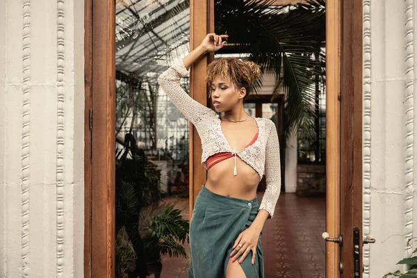 Mujer afroamericana joven de moda en punto superior y falda posando cerca de la entrada del jardín interior en el fondo, mujer de moda disfrutando de vibraciones de verano — Stock Photo