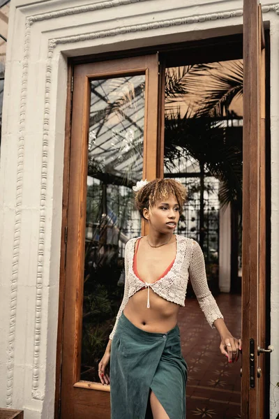 Mulher americana africana jovem confiante em top de malha e roupa de verão olhando para longe enquanto estava perto da porta aberta do jardim interior, mulher elegante desfrutando de vibrações de verão — Fotografia de Stock