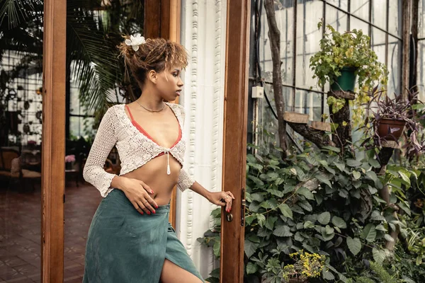 Seitenansicht der stilvollen jungen afrikanisch-amerikanischen Frau im Sommer-Outfit und Strickoberteil Tür des Indoor-Gartens im Hintergrund, modische Frau genießt sommerliche Stimmung — Stockfoto