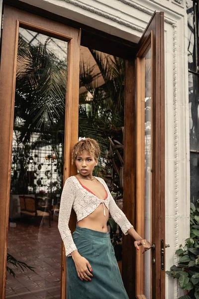 Junge afrikanisch-amerikanische Frau im Sommer-Outfit und Stricktop öffnet Tür des Indoor-Gartens und schaut weg, modische Frau genießt sommerliche Stimmung, botanische — Stockfoto
