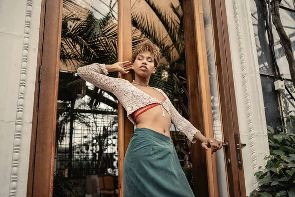 Vista de ángulo bajo de la joven mujer afroamericana de moda en traje de verano de pie cerca de la puerta del jardín interior en el fondo, mujer de moda disfrutando de vibraciones de verano - foto de stock