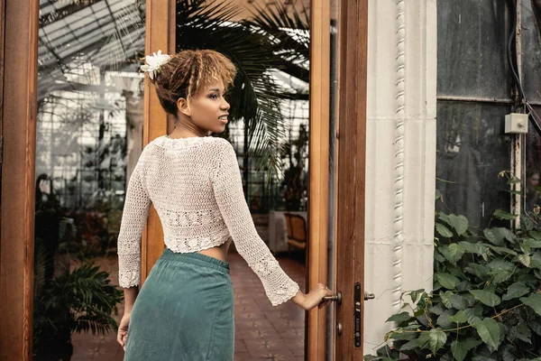 Joven mujer afroamericana alegre en traje de verano mirando hacia otro lado mientras está de pie cerca de la puerta del jardín interior en el fondo, señora elegante rodeada de follaje tropical exótico - foto de stock