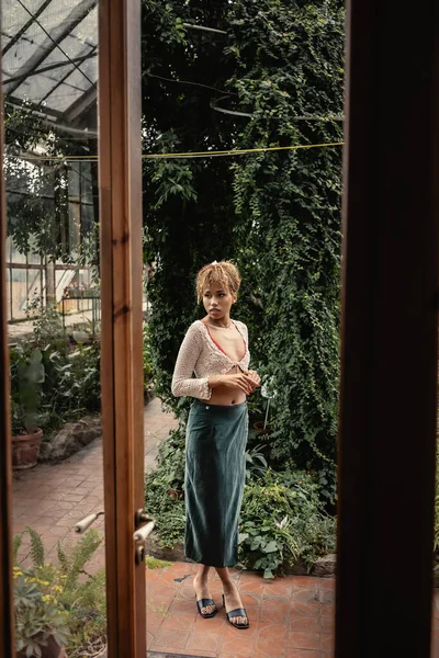 Pleine longueur de jeune femme afro-américaine à la mode en tricot haut et jupe debout près des plantes et porte du jardin intérieur, dame élégante entourée de feuillage tropical exotique, concept d'été — Photo de stock