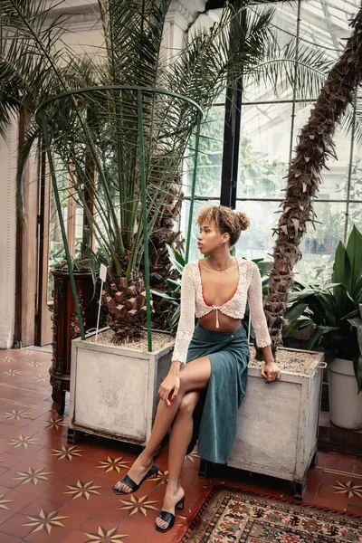 Volle Länge von trendigen jungen afrikanisch-amerikanischen Frau im Sommer-Outfit wegschauen, während sie in der Nähe von Palmen im Garten sitzen, Fashionista posiert inmitten tropischer Flora, Sommerkonzept — Stockfoto