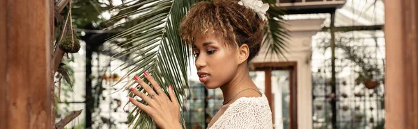 Young and stylish african american woman in knitted top touching brunch of palm tree and standing in blurred garden center at background, fashionista posing amidst tropical flora, banner — Stock Photo