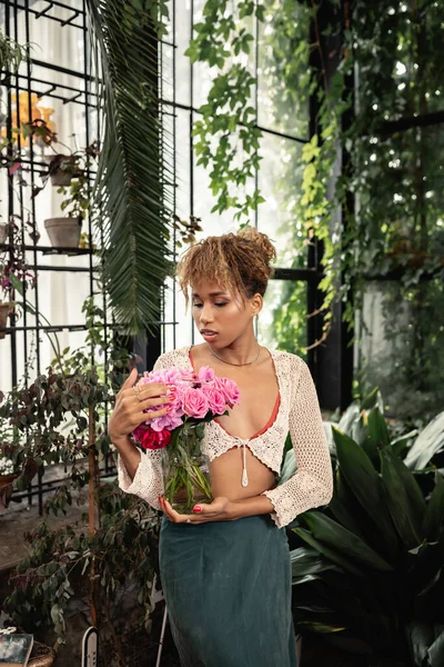 Mujer afroamericana joven de moda en jarrón de punto superior con rosas y de pie cerca de plantas borrosas en el centro del jardín en el fondo, mujer de moda con estilo tropical, concepto de verano - foto de stock
