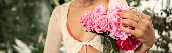 Обрізаний вид розмитої молодої афроамериканки в в'язаному верхньому тримає вазу з рожевими трояндами в розмитому критому саду на фоні, модна жінка з тропічним підносом, банер — стокове фото
